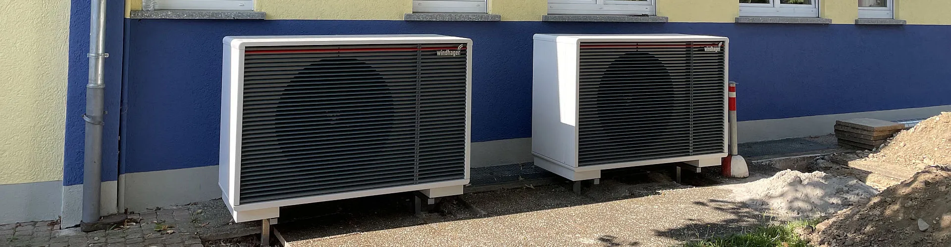 Zwei Wärmepumpen-Außeneinheiten von Windhager stehen vor einem blau-gelben Gebäude.
