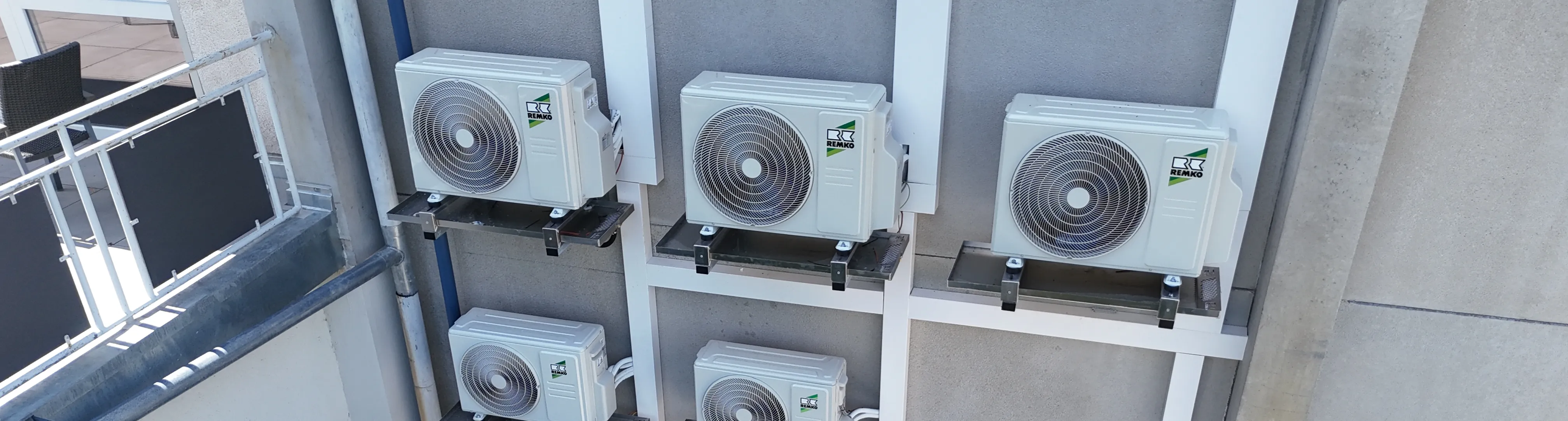 Sechs weiße Außengeräte von Klimaanlagen der Marke Remko, montiert an einer Hauswand auf Metallhalterungen.