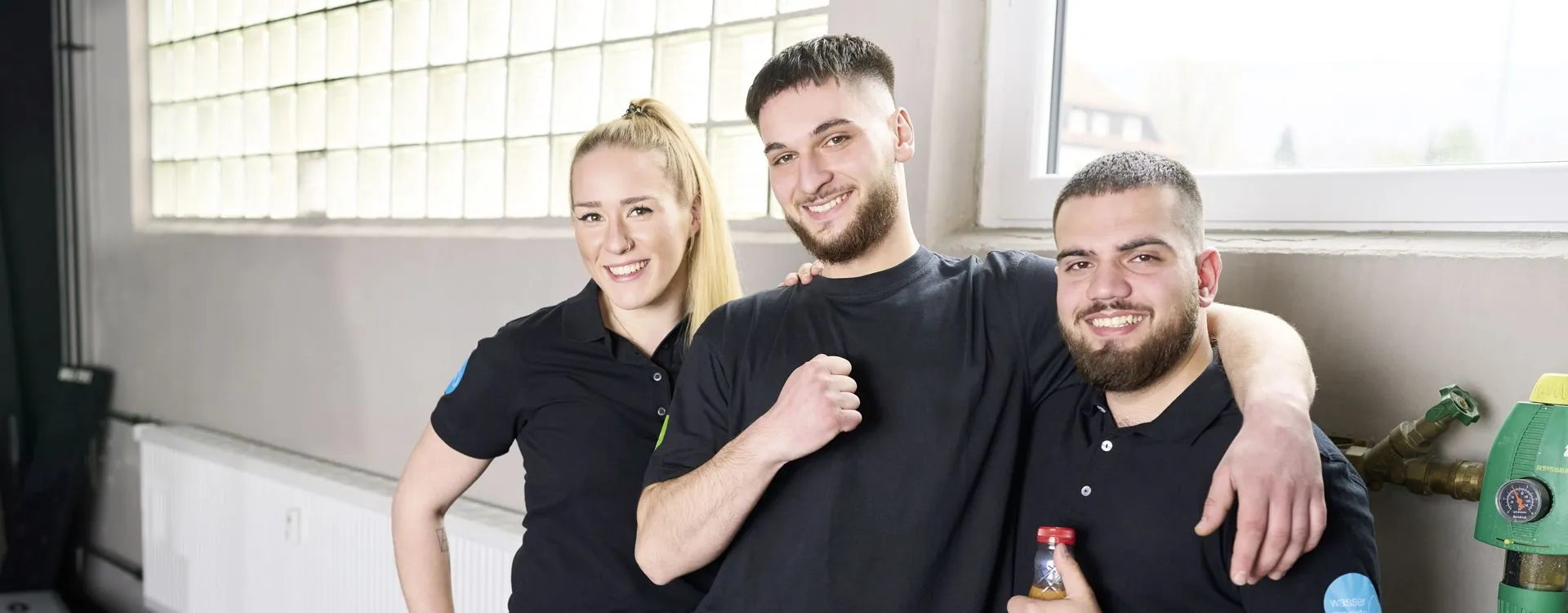 Drei junge Auszubildende Anlagenmechaniker posieren in Arbeitskleidung vor dem Werkstattsfenster.