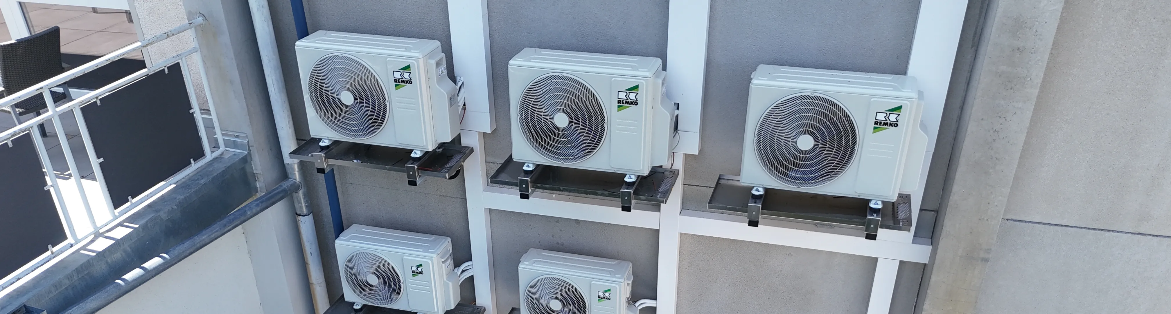 Mehrere außen montierte Klimageräte und Wärmepumpen in zwei Reihen an einer Gebäudewand, befestigt auf Metallkonsolen, mit Rohren und Kabeln, die nach unten geführt sind; daneben ein kleiner Hofbereich und eine angelehnte Leiter.