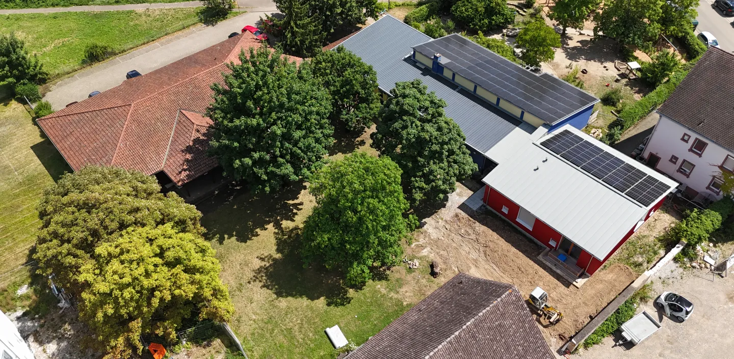 Luftaufnahme eines Gebäudekomplexes mit mehreren Häusern und Bäumen. In der Mitte befindet sich eine Grünfläche mit großen Bäumen. Rechts sind zwei moderne Gebäude mit grauen Dächern und Solaranlagen zu sehen, eines davon rot gestrichen.