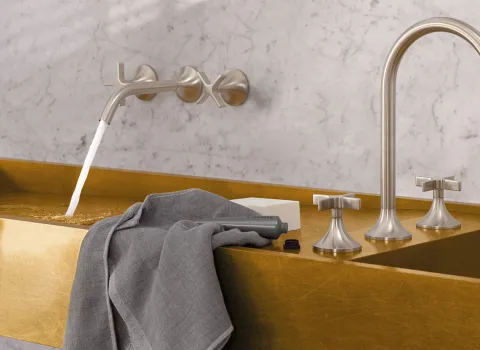 Moderne Armatur mit Wasserstrahl in ein goldenes Becken. Daneben ein graues Handtuch und eine Pflegecremetube.