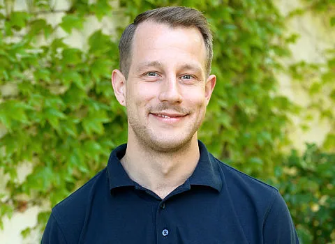 Ein Mitarbeiter der Kreuz GmbH im dunklen Hemd steht vor einer begrünten Mauer und lächelt freundlich in die Kamera.