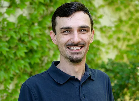 Ein junger Mitarbeiter in dunklem Hemd lächelt in die Kamera.