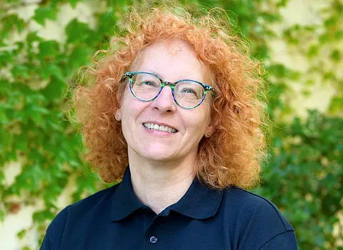 Portrait einer lächelnden Frau im dunklen Hemd vor grün bewachsener Wand.