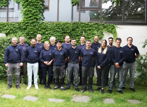 Vor einer mit Efeu bewachsenen Wand steht eine Gruppe von Menschen in dunkelblauer Teamkleidung nebeneinander.