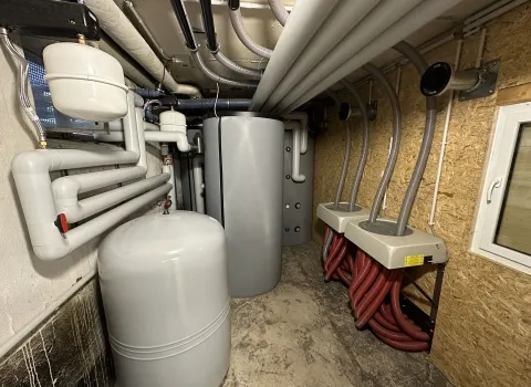 In einem Technikraum befinden sich ein großer grauer Pufferspeicher und ein weiterer runder Druckbehälter, die Teil einer komplexen Heiztechnik sind. 