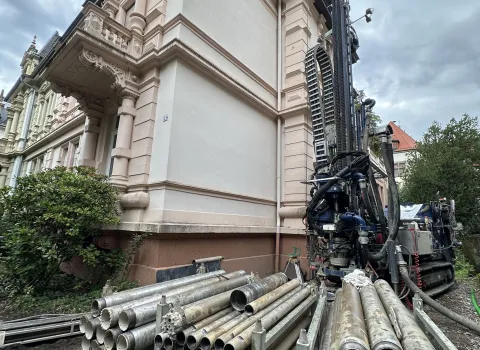 Baustelle an einem historischen Gebäude mit Bohrgerät und gestapelten Metallrohren im Vordergrund, Vorbereitung für energetische Sanierungsmaßnahmen.