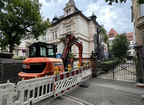 Baustellenszene vor einem historischen Gebäude: Ein orangefarbener Bagger hinter Absperrgittern im Einsatz für Sanierungs- oder Erdbohrarbeiten.