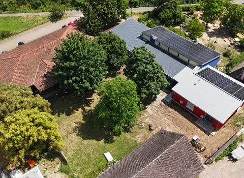 Luftaufnahme eines Gebäudekomplexes mit mehreren Häusern und Bäumen. In der Mitte befindet sich eine Grünfläche mit großen Bäumen. Rechts sind zwei moderne Gebäude mit grauen Dächern und Solaranlagen zu sehen, eines davon rot gestrichen.