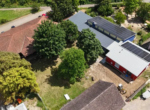 Luftaufnahme eines Gebäudekomplexes mit mehreren Häusern und Bäumen. In der Mitte befindet sich eine Grünfläche mit großen Bäumen. Rechts sind zwei moderne Gebäude mit grauen Dächern und Solaranlagen zu sehen, eines davon rot gestrichen.