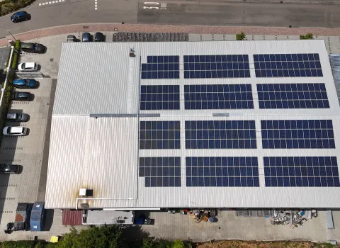 Industrie- oder Geschäftsgebäude mit hellgrauem Metalldach, auf dem eine große Photovoltaikanlage installiert ist. Die Solarmodule sind in mehreren Reihen angeordnet, davor Parkplätze mit geparkten Autos und eine angrenzende Straße.