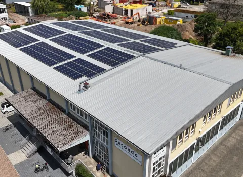 Großes Gewerbegebäude mit hellgrauem Blechdach, auf dem zahlreiche Solarmodule installiert sind; davor ein Eingangsbereich mit überdachtem Zugang, umliegende Parkflächen und im Hintergrund eine Baustelle mit Maschinen und Materialien.