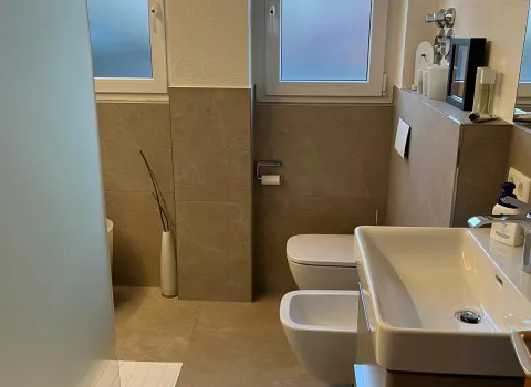 Badezimmer-Referenz mit Waschtisch, Bidet und Waschbecken auf grauen Fliesen vor zwei Fenstern.