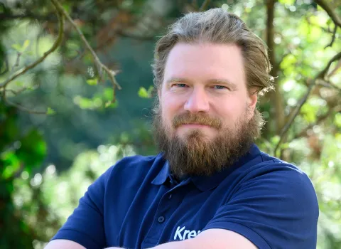 Portrait einer lächelnden Frau im dunklen Hemd vor grünen Blättern.