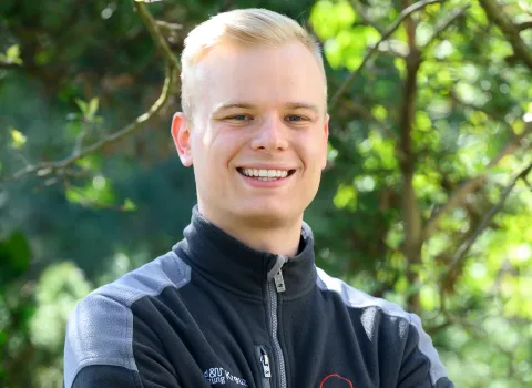 Portrait eines lächelnden jungen Mannes im dunklen Hemd vor grünen Blättern.
