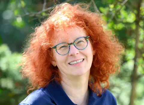 Portrait einer lächelnden Frau im dunklen Hemd vor grünen Blättern.