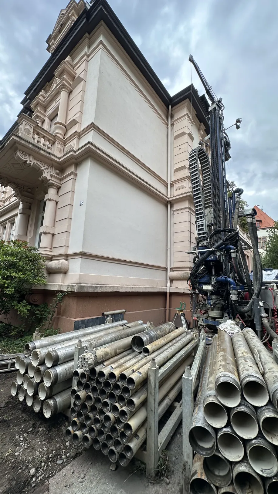 Baustelle an einem historischen Gebäude mit Bohrgerät und gestapelten Metallrohren im Vordergrund, Vorbereitung für energetische Sanierungsmaßnahmen.