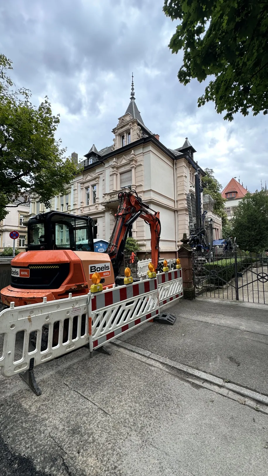 Baustellenszene vor einem historischen Gebäude: Ein orangefarbener Bagger hinter Absperrgittern im Einsatz für Sanierungs- oder Erdbohrarbeiten.