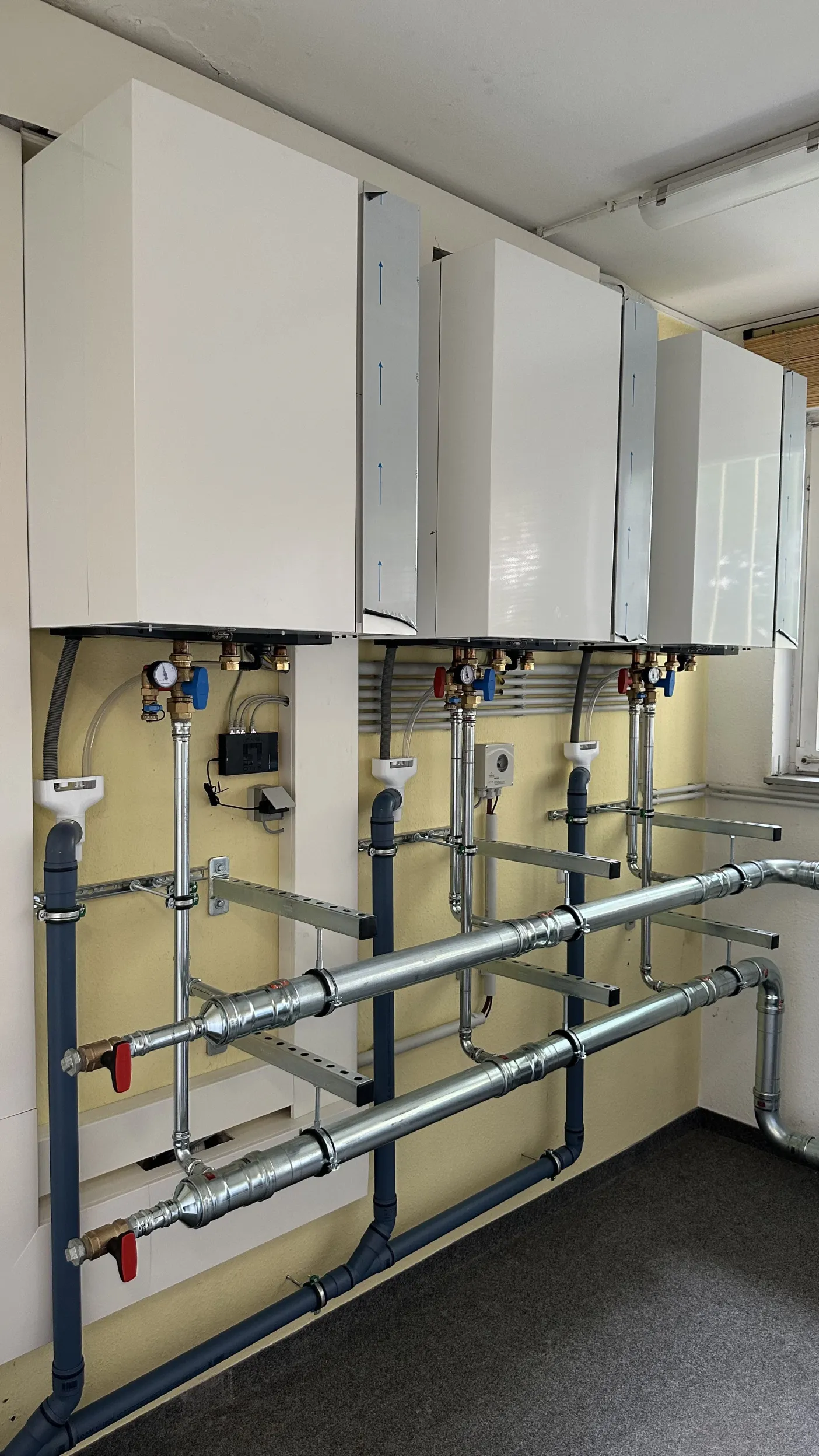 Drei weiße Wandgeräte in Reihe, darunter verzinkte Sammelleitungen, senkrechte Rohrstränge und Absperrarmaturen. Einige Bauteile haben noch Schutzfolien. Rechts befindet sich ein Fenster, links sind Montageprofile an der Wand sichtbar.