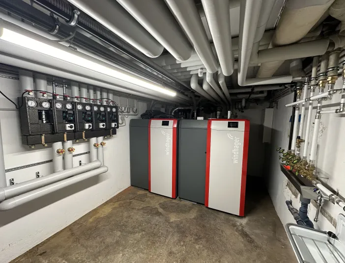 In einem großen, gut beleuchteten Technikraum stehen zwei moderne Windhager-Heizsysteme in Rot und Grau. 
