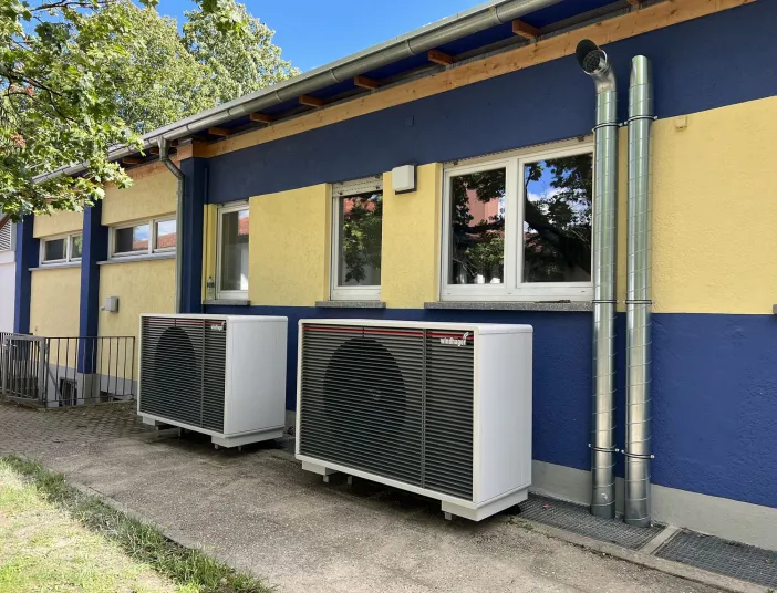 Zwei moderne Wärmepumpen sind nebeneinander an einer blau-gelb gestrichenen Außenwand eines Gebäudes installiert.