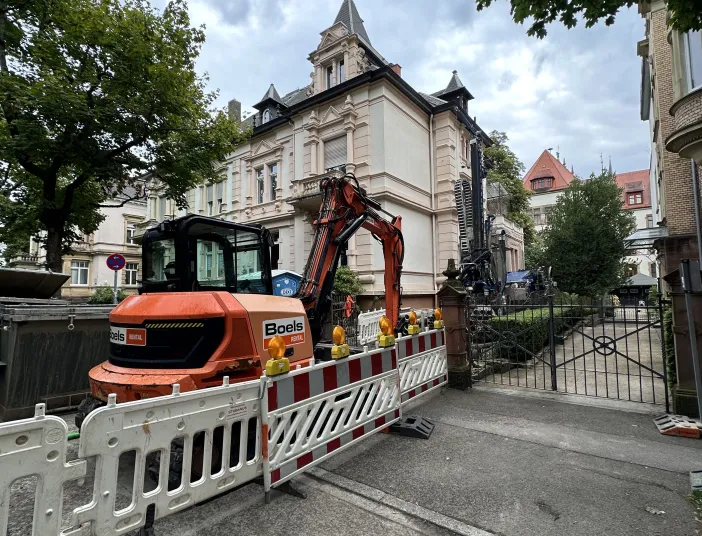 Baustellenszene vor einem historischen Gebäude: Ein orangefarbener Bagger hinter Absperrgittern im Einsatz für Sanierungs- oder Erdbohrarbeiten.