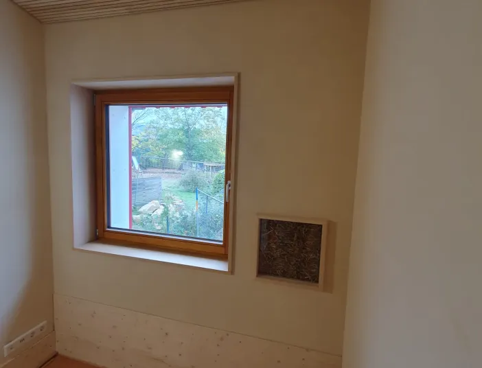 Innenaufnahme eines hellen Raums mit Holzdecke und Holzfußboden. In der Mitte ist ein Fenster mit Holzrahmen zu sehen, durch das man auf einen Garten blickt. Rechts neben dem Fenster befindet sich ein kleines Wandfenster mit sichtbarer Stroh- oder Naturdämmung als Baumaterialprobe.