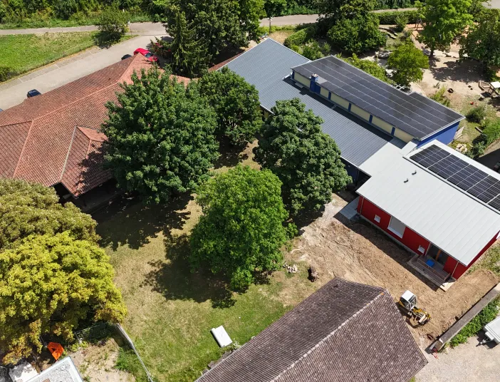 Luftaufnahme eines Gebäudekomplexes mit mehreren Häusern und Bäumen. In der Mitte befindet sich eine Grünfläche mit großen Bäumen. Rechts sind zwei moderne Gebäude mit grauen Dächern und Solaranlagen zu sehen, eines davon rot gestrichen.