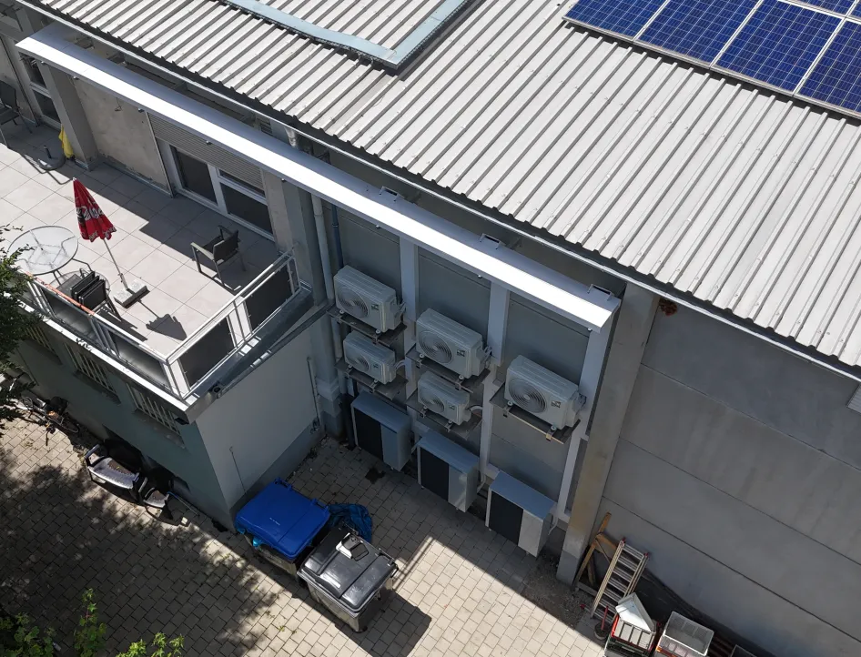 Gebäudeansicht mit montierten Klimageräten an der Außenwand, jeweils in zwei Reihen übereinander angebracht. Rechts ist das Metalldach mit Solarpaneelen zu sehen, links ein Balkon mit Sitzmöbeln und Sonnenschirm.