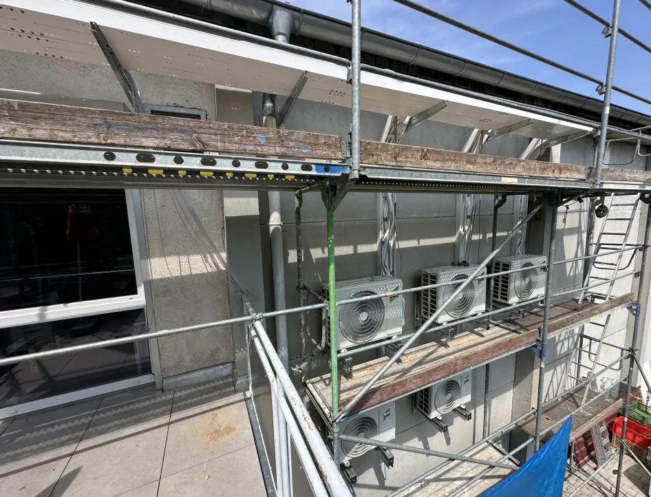 Mehrere Klimageräte der Marke Remko sind an der Fassade eines Gebäudes montiert, davor steht ein Baugerüst. Kabel und Leitungen führen von den Geräten nach oben unter das Dach.