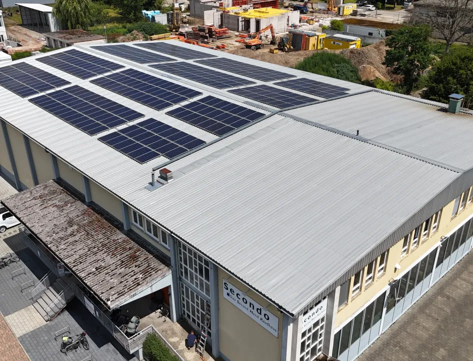 Großes Gewerbegebäude mit hellgrauem Blechdach, auf dem zahlreiche Solarmodule installiert sind; davor ein Eingangsbereich mit überdachtem Zugang, umliegende Parkflächen und im Hintergrund eine Baustelle mit Maschinen und Materialien.